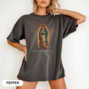 May include: Dark grey t-shirt with an oval illustration of the Virgin Mary, wearing a blue cloak and pink dress. The text "pray like a mom" is below the illustration. The word "PEPPER" is at the bottom of the shirt.