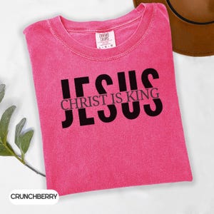 A pink t-shirt with the text "JESUS IS KING" printed on it, along with a brown hat and some greenery in the background.