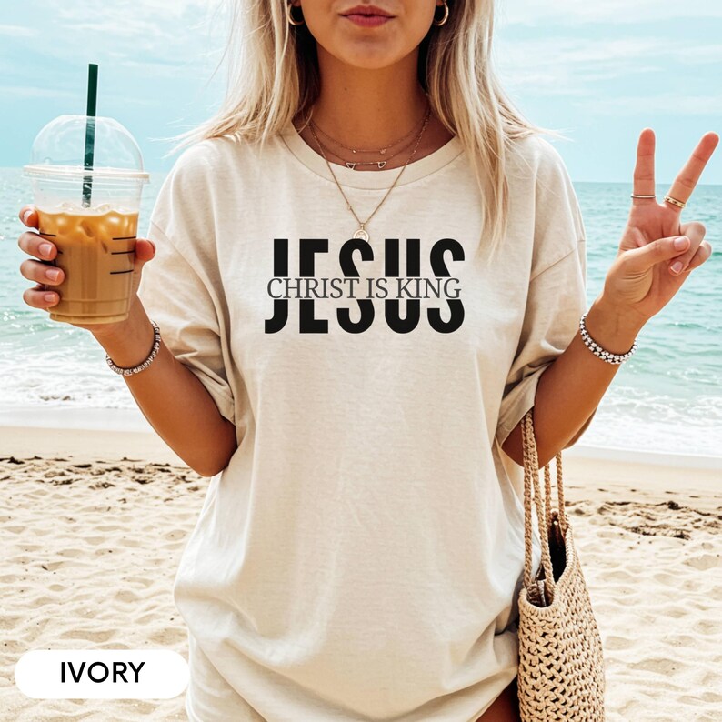 A woman wearing a white t-shirt with the text "JESUS CHRIST IS KING" printed on it, standing on a beach with her hands in a peace sign. She is holding a drink in a plastic cup with a straw.