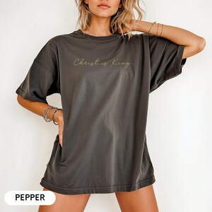 A woman wearing a gray t-shirt with the text "Christ is King" printed on it, posing with her hand on her hip.