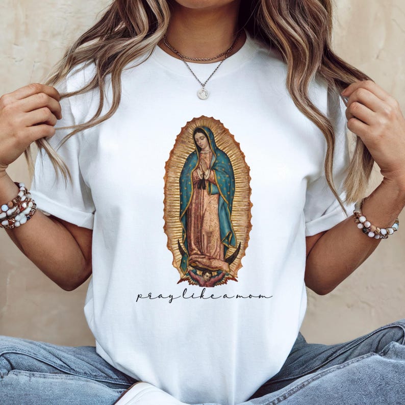 May include: White t-shirt with a religious image of a woman in a blue and gold halo. The image is centered on the shirt, with the words "pray like a mom" in a script font below the image. The shirt is made of a soft material.