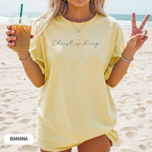A woman wearing a yellow t-shirt with the text "Christ is King" printed on it, standing on a beach and holding a drink in her hand.