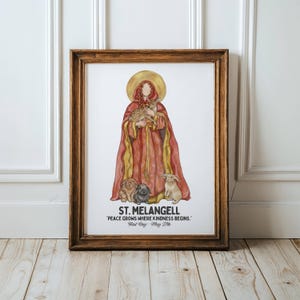 May include: Framed watercolor print featuring St. Melangell, holding a fawn, surrounded by rabbits. The saint is depicted in a red and gold robe with a halo. Text at the bottom reads "ST. MELANGELL PEACE GROWS WHERE KINDNESS BEGINS. Feast Day: May 27th."