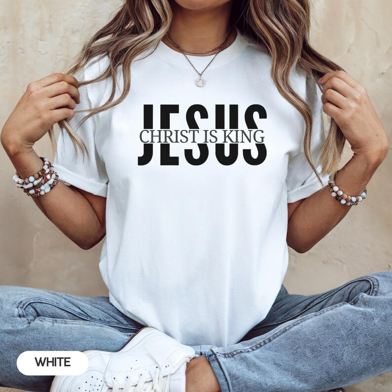 A woman wearing a white t-shirt with the text "JESUS IS KING" printed on it, posing with her hands behind her back.