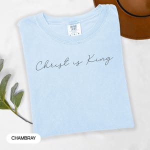 A light blue t-shirt with the text "Christ is King" printed on it, along with a brown hat and some greenery in the background.
