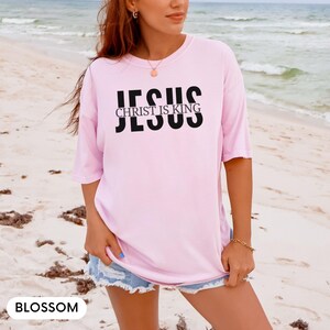 A woman wearing a pink t-shirt with the text "JESUS IS KING" printed on it, standing on a beach.