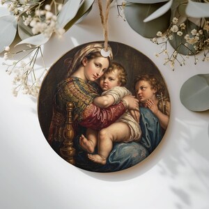 Puede incluir: Un adorno redondo de cerámica con una pintura al óleo clásica de una mujer con pañuelo estampado y prenda roja sosteniendo a un niño pequeño, con otro niño cerca. El adorno cuelga de un cordel rústico sobre un fondo blanco.