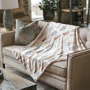 May include: A cream-colored throw blanket featuring a repeating pattern of various religious figures in blue, red, and gold robes, interspersed with small blue fleur-de-lis symbols, draped over a beige upholstered sofa with a dark patterned pillow.