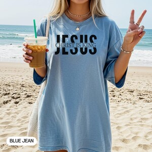 A woman wearing a blue t-shirt with the text "JESUS IS KING" is standing on a beach, holding a drink and making a peace sign.