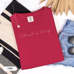 a red t-shirt with the text "Christ is King" printed on it, along with a pair of denim shorts and sunglasses.