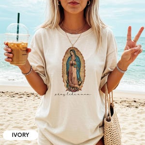 May include: Ivory t-shirt with a religious illustration of a saint in a gold oval frame. The saint is wearing blue and red robes. The text "pray like a mom" is printed below the illustration. The shirt is worn on a beach.