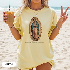 May include: A pale yellow t-shirt featuring an oval graphic of the Virgin Mary. The text "pray like a mom" is written below the image. The word "BANANA" is printed at the bottom left. The t-shirt is worn on a beach.