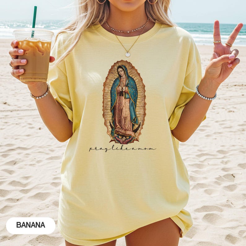May include: A pale yellow t-shirt featuring an oval graphic of the Virgin Mary. The text "pray like a mom" is written below the image. The word "BANANA" is printed at the bottom left. The t-shirt is worn on a beach.