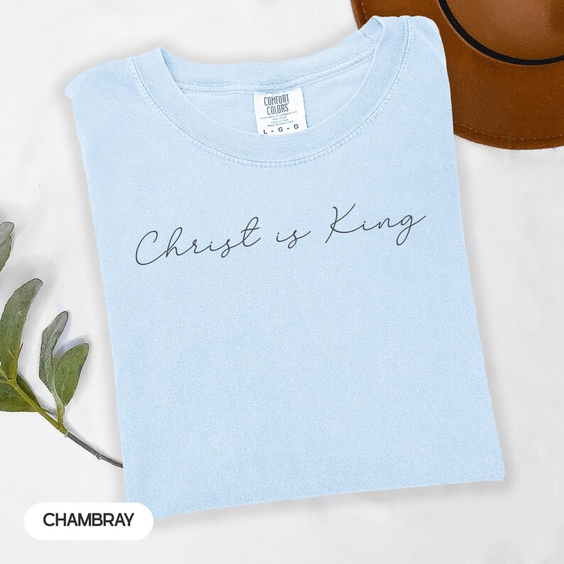 A light blue t-shirt with the text "Christ is King" printed on it, along with a brown hat and some greenery in the background.