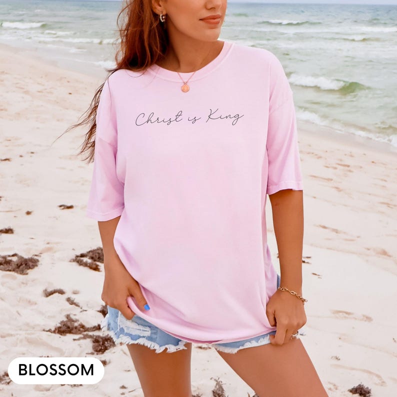 A woman wearing a pink t-shirt with the text "Christ is King" standing on a beach with the ocean in the background.