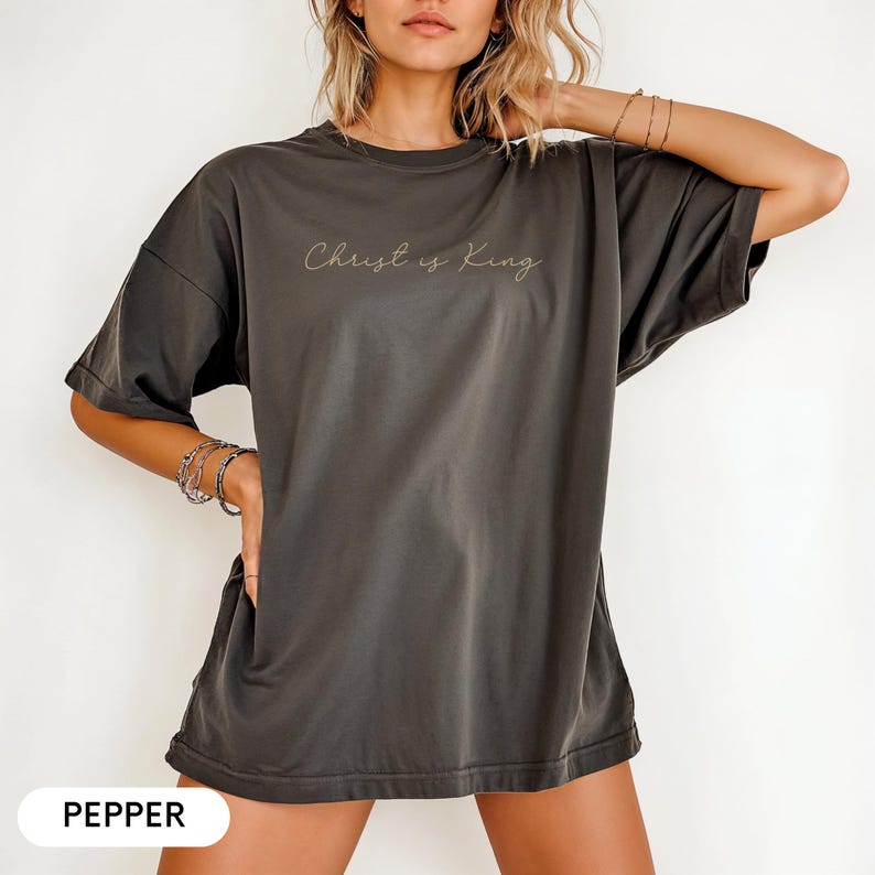 A woman wearing a gray t-shirt with the text "Christ is King" printed on it, posing with her hand on her hip.