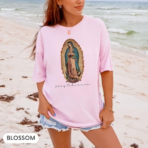 May include: A pale pink t-shirt with an image of the Virgin Mary in a gold frame and the words "pray like a mom." The shirt is made of a soft fabric.