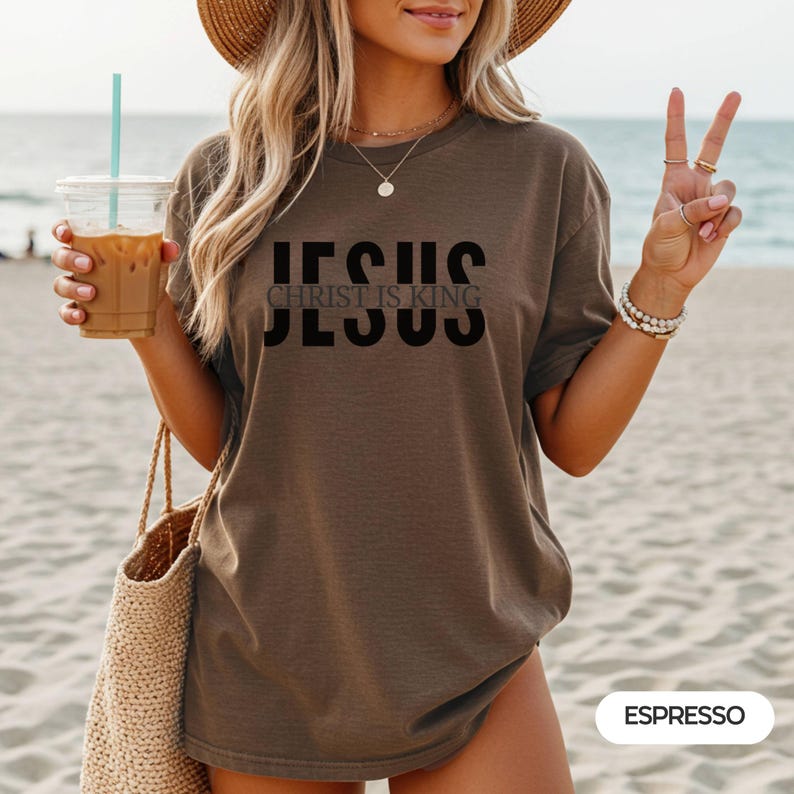 A woman wearing a brown t-shirt with the text "JESUS IS KING" and holding a drink, standing on a beach.