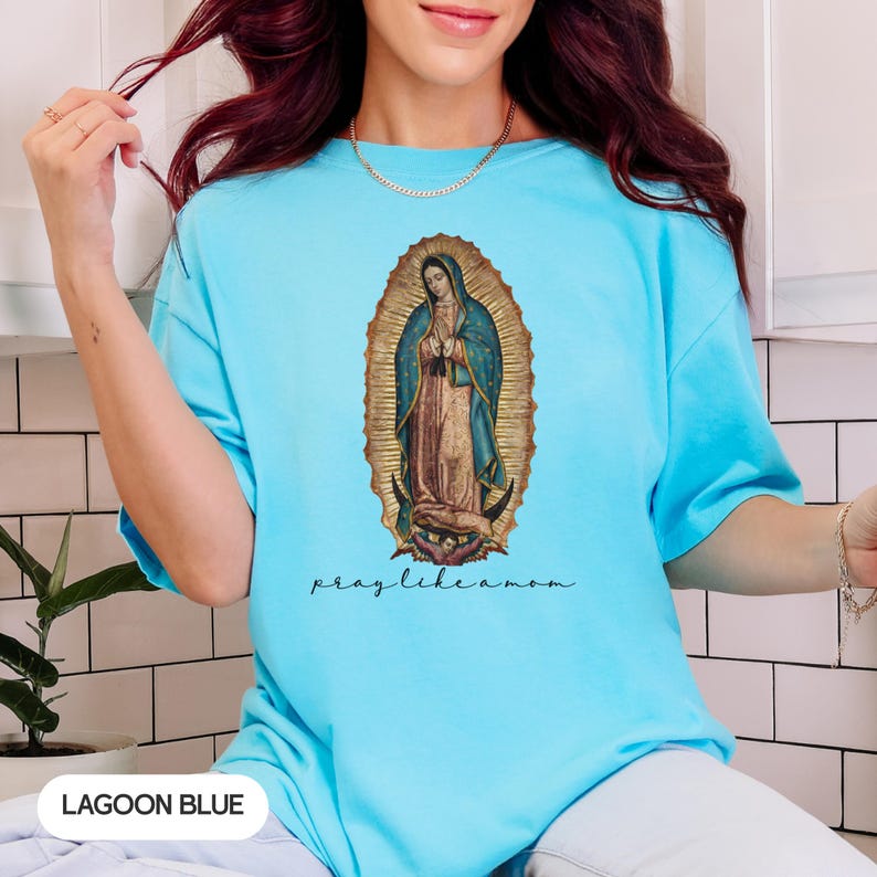 May include: Lagoon blue t-shirt with an image of the Virgin Mary in a gold oval frame. The words "pray like a mom" are written in a script font below the image. The shirt is a solid colour.