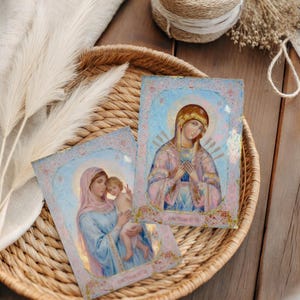 May include: Two religious icon prints featuring the Virgin Mary and child, displayed in a woven basket. The icons have pastel colors, with gold accents and text. The basket sits on a wooden surface, with decorative elements.