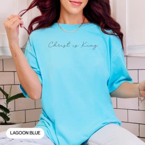 A woman with long, dark hair wearing a light blue t-shirt with the text "Christ is King" printed on it.
