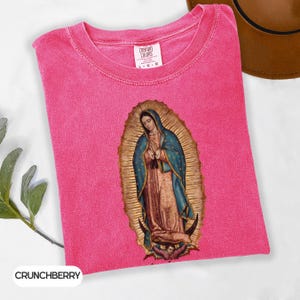 May include: A vibrant pink t-shirt with a crew neck and short sleeves. The shirt displays an image of the Virgin Mary in a gold halo, wearing a blue cloak and a tan dress. The shirt is labelled "Comfort Colors" and "Crunchberry".