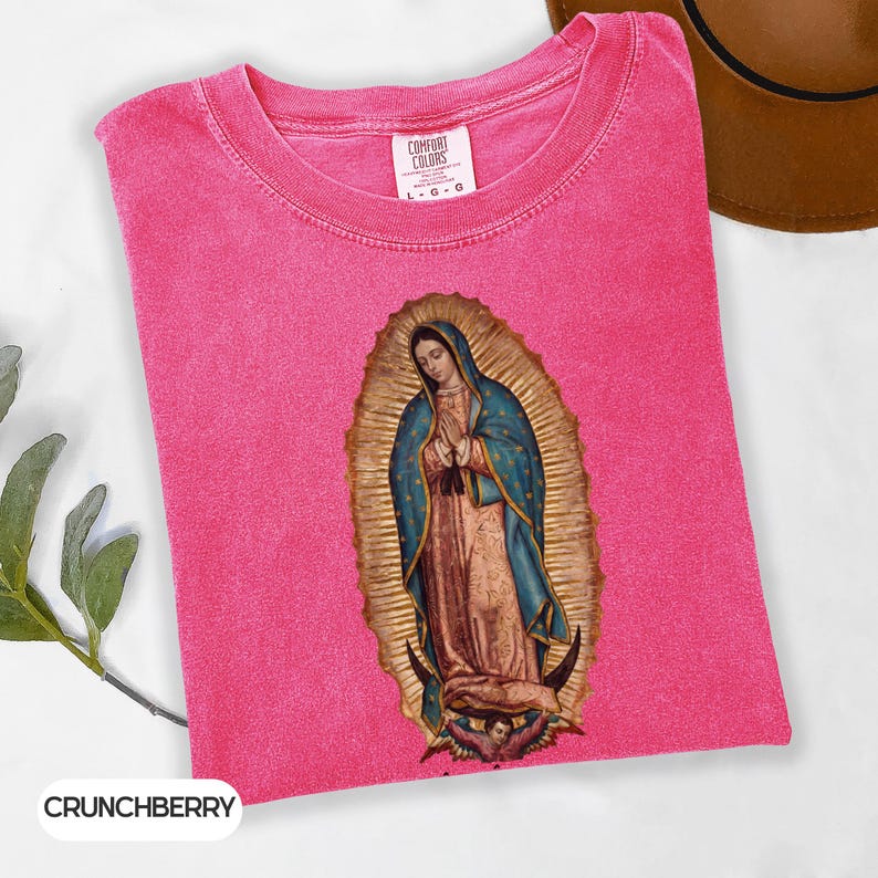May include: A vibrant pink t-shirt with a crew neck and short sleeves. The shirt displays an image of the Virgin Mary in a gold halo, wearing a blue cloak and a tan dress. The shirt is labelled "Comfort Colors" and "Crunchberry".