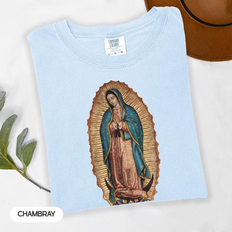 May include: Pale blue t-shirt featuring a graphic of Our Lady of Guadalupe. The image shows a detailed illustration of the Virgin Mary, with a gold halo and blue and pink robes. The shirt is labelled "Chambray".