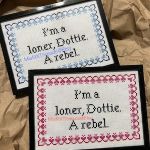 May include: Two framed cross-stitch embroidery pieces with a white background and black text. The top piece has a blue and white border and the bottom piece has a pink and white border. Both pieces say "I'm a loner, Dottie. A rebel."
