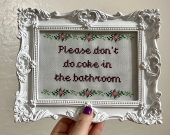 May include: White framed cross-stitch art with the text "Please don't do coke in the bathroom." The text is in dark red, with floral accents in green and pink. The frame has ornate detailing.