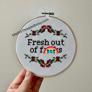 May include: A white embroidery hoop with a cross-stitch design that reads "Fresh out of f***s" with a rainbow and a crying face emoji. The hoop is surrounded by a floral border of red flowers and green leaves.