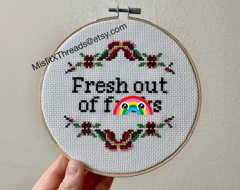 May include: A white embroidery hoop with a cross-stitch design that reads "Fresh out of f***s" with a rainbow and a crying face emoji. The hoop is surrounded by a floral border of red flowers and green leaves.