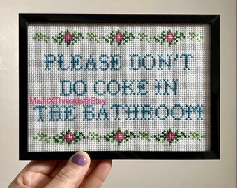 May include: A framed cross-stitch with a white background and blue text that reads "Please don't do coke in the bathroom". The text is surrounded by a green vine with pink flowers.