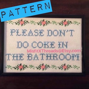 May include: A cross-stitch pattern with a black frame and a white background. The pattern reads "Please don't do coke in the bathroom." with pink flowers along the top and bottom.