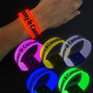 May include: A collection of glowing wristbands in various colors, including orange, pink, yellow, white, blue, and green. Each band features the text "Jimmy & Camelia" in black. The wristbands are illuminated, creating a vibrant and eye-catching display.