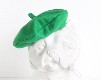 May include: A white ceramic dog figurine wearing a green beret. The beret is made of felt and has a simple design.