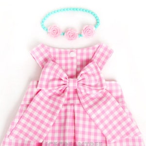 Puede incluir: Un vestido de cuadros vichy rosa y blanco con un gran lazo rosa en la espalda. El vestido es sin mangas y tiene una falda fruncida. Se incluye una diadema azul claro con detalles de flores rosas.
