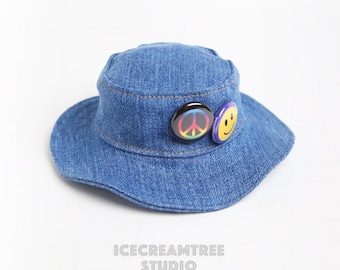 May include: A blue denim bucket hat with a peace sign and smiley face pin. The hat has a wide brim and a flat crown.