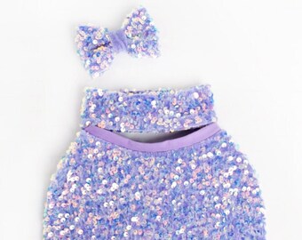 May include: A purple sequined baby outfit with a matching purple bow. The outfit has a halter top with a cutout in the front and a skirt.