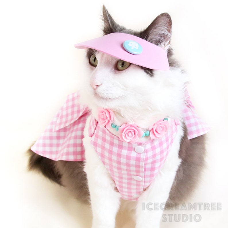 Puede incluir: Un gato blanco y gris con un vestido de cuadros rosa y blanco, una visera rosa y un collar de flores rosas. El gato est&aacute; mirando hacia la izquierda de la imagen. El texto "ICECREAMTREE STUDIO" est&aacute; en la esquina inferior derecha de la imagen.