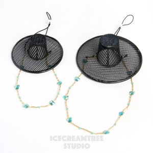 May include: Two black mesh hats with gold chains and turquoise beads hanging from them. The hats are hanging from a white background.