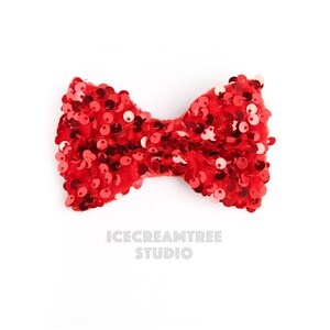 May include: A bright red bow with a velvet texture and covered in shiny sequins. The bow is centered on a white background. The text "ICECREAMTREE STUDIO" is printed below the bow.