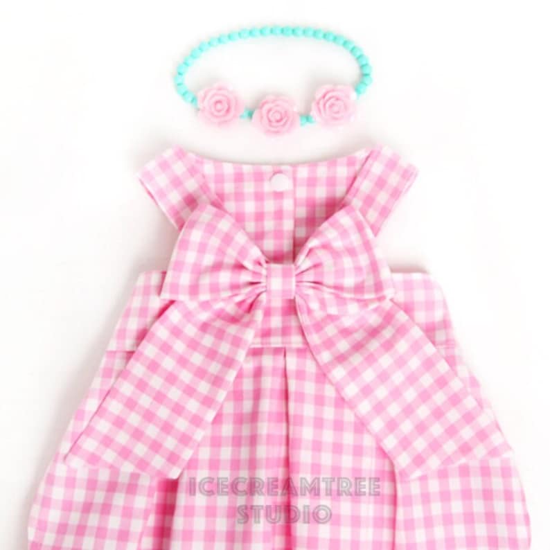 Puede incluir: Un vestido de cuadros vichy rosa y blanco con un gran lazo rosa en la espalda. El vestido es sin mangas y tiene una falda fruncida. Se incluye una diadema azul claro con detalles de flores rosas.