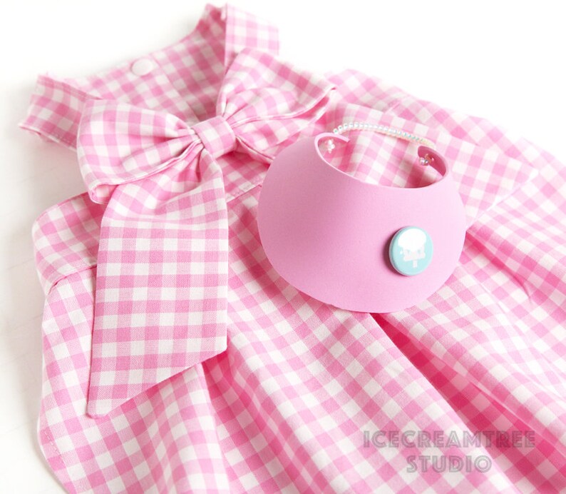 Puede incluir: Vestido de perro de cuadros vichy rosa y blanco con un gran lazo rosa y un collar rosa con un dise&ntilde;o de nube blanca.