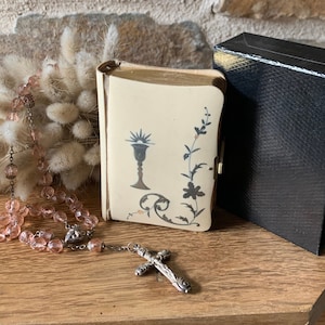 May include: A cream-coloured, book-shaped container with a floral and chalice design, a black box, and a pink beaded rosary with a silver cross rest on a wooden surface. The background features a stone wall and dried flowers.