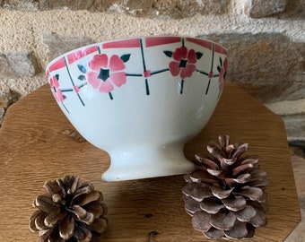 May include: A white ceramic bowl with a pink floral design around the rim. The bowl sits on a wooden surface with two pine cones. The bowl has a pedestal base and a slightly flared shape.