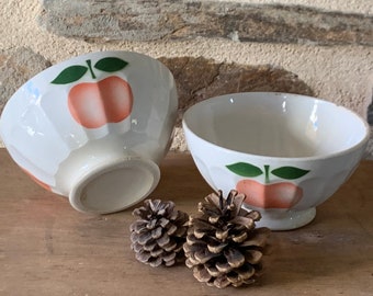 May include: Two white ceramic bowls with a peach design. The bowls have a fluted edge and a white base. The peaches are pink with green leaves. The bowls are sitting on a wooden table.