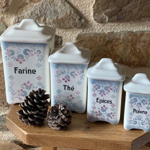 Puede incluir: Cuatro botes de cerámica blancos con tapas, etiquetados como "Farine", "Thé", "Epices" y "Poivre". Cada bote presenta un diseño floral azul y rosa. Dos piñas se encuentran frente a los botes sobre una superficie de madera.
