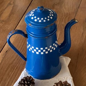 Op de afbeelding: Blauwe emaille koffiepot met een wit geruit ontwerp. De pot heeft een deksel en een tuit. Hij staat op een witte doek met dennenappels.