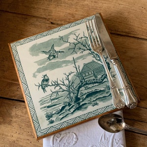 May include: A square ceramic tile with a green and white design of birds and a small house. The tile is framed in wood and is sitting on a wooden table. A white linen napkin with an embroidered monogram is next to the tile.